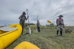 Three volunteers standing alongside two yellow canoes on the shore, preparing for a community water activity.