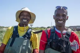Two smiling men wearing waders, standing in the Khayelitsha Wetlands, with blue skies in the background.
