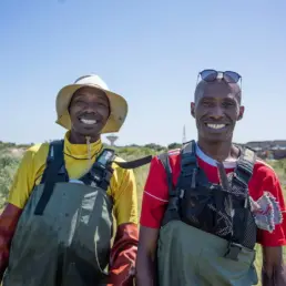 Two smiling men wearing waders, standing in the Khayelitsha Wetlands, with blue skies in the background.
