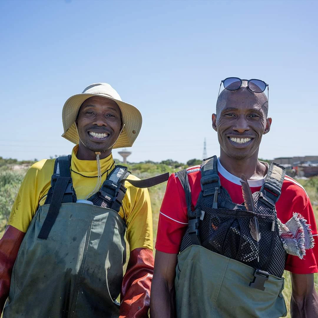 Two smiling men wearing waders, standing in the Khayelitsha Wetlands, with blue skies in the background.